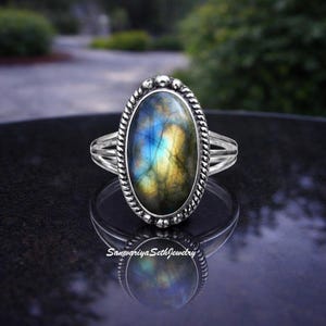 May include: Oval-shaped silver ring featuring a large, iridescent labradorite stone with blue, yellow, and green hues. The stone is set in a detailed bezel with a twisted rope design. The ring has a split shank.