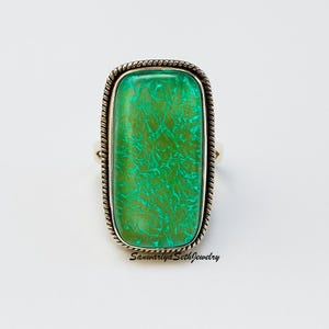 May include: A rectangular ring featuring a vibrant green stone with a unique pattern. The stone is set in a silver-toned metal with a rope-like border. The ring has a simple band and the text "Sanwariya SethJewelry" is visible.
