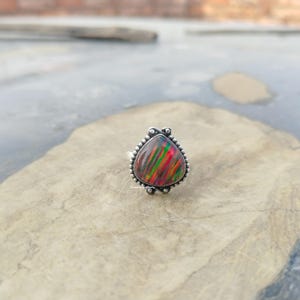 Op de afbeelding: Zilveren ring met een driehoekige zwarte opaal met rode, groene en gele strepen. De opaal is gezet in een kralenrand. De ring wordt gepresenteerd op een lichtgekleurd stenen oppervlak.