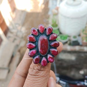 May include: A silver-toned ring featuring a large, oval-shaped, deep red gemstone at the center, surrounded by smaller, teardrop-shaped red stones. The ring has a floral-like design, with the gemstones set against a dark background. The ring is a piece of jewelry.
