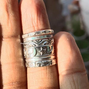 Könnte beinhalten: Eine Ansammlung von Silberringen, die auf einem Finger gestapelt sind. Die Ringe weisen verschiedene Designs auf, darunter strukturierte Bänder, wirbelnde Muster und Himmelsmotive wie Monde und Sterne. Die Ringe werden im natürlichen Licht präsentiert.