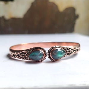 Puede incluir: Brazalete de puño de color cobre con piedras preciosas verdes iridiscentes en forma de lágrima. El brazalete presenta diseños ornamentados y arremolinados alrededor de las piedras preciosas y a los lados. El diseño abierto facilita su uso.