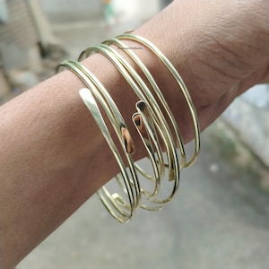 Op de afbeelding: Een set goudkleurige armbanden om een pols. De armbanden zijn dun, gebogen en hebben een spiraalvormig ontwerp. De armbanden zijn gemaakt van glanzend metaal en worden samen gedragen. De algehele uitstraling is elegant en minimalistisch.