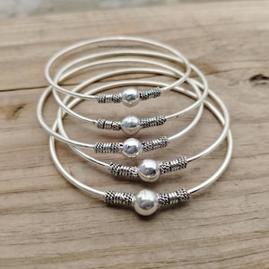 Könnte beinhalten: Fünf silberne Armreifen mit einer zentralen silbernen Perle und dekorativen Details. Die Armreifen werden auf einer Holzoberfläche präsentiert. Der Text "SanwariyaSethJewelry" ist im Hintergrund sichtbar.