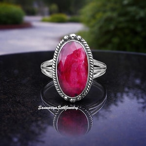 Könnte beinhalten: Ein Silberring mit einem ovalen, facettierten, tiefroten Edelstein. Der Edelstein ist in einer strukturierten, seilartigen Lünette gefasst. Der Ring hat ein geteiltes Schaftdesign. Der Ring wird auf einer reflektierenden schwarzen Oberfläche präsentiert.