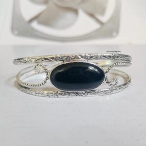 Può includere: Bracciale color argento con una pietra nera ovale. Il bracciale presenta un motivo floreale lungo i bordi e un ricciolo decorativo su ciascun lato della pietra. È un gioiello.