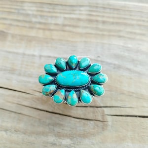 May include: A silver ring featuring a large oval turquoise stone surrounded by teardrop-shaped turquoise stones. The ring is set against a wooden background, showcasing the vibrant blue-green hues of the gemstones.