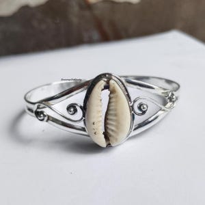 Può includere: Un bracciale rigido argentato con un elemento centrale a forma di conchiglia color crema. Il bracciale presenta un design delicato e aperto con volute e curve decorative. La conchiglia è il punto focale, aggiungendo un elemento naturale al gioiello.