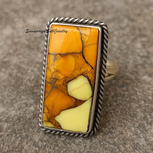 May include: A rectangular silver ring featuring a large, eye-catching stone. The stone is a vibrant mix of yellow and orange hues with a unique, cracked pattern. The ring has a twisted rope-like border and the text "Sanwariya Seth Jewelry".