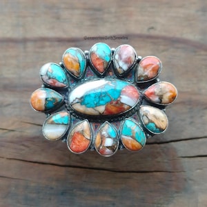 以下が含まれることがあります： 中央にターコイズと白の大きな楕円形の石を配し、ターコイズ、オレンジ、白の涙型の石で囲まれたシルバーリング。木製の背景にセットされており、複雑なデザインが特徴です。