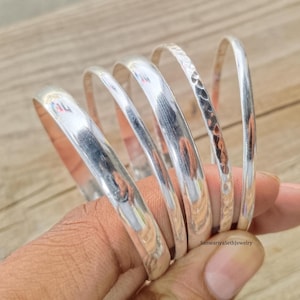 Peut inclure: Une main tient une pile de bracelets argentés. Les bracelets ont une surface polie et réfléchissante, dont l'un présente un motif texturé martelé. Les bijoux sont présentés sur un fond en bois brun clair.