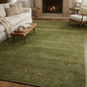 May include: A large, rectangular green rug with a textured surface. The rug is the focal point of a living room, with a white sofa, wooden coffee table, and fireplace in the background. The rug has small, decorative accents.