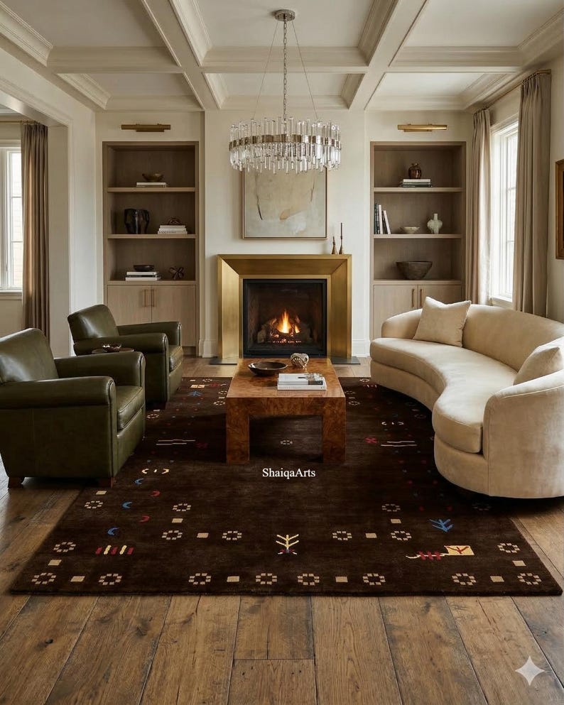 May include: A living room with a brown patterned rug, a curved beige sofa, and two green leather armchairs. A fireplace with a gold frame and a crystal chandelier add elegance. The room has wooden floors and built-in bookshelves.