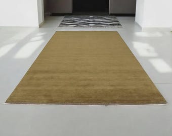 May include: A rectangular, golden-brown rug dominates the foreground, its texture subtly visible. In the background, a smaller black and white patterned rug adds contrast. The setting is a bright, minimalist interior with white walls and flooring.