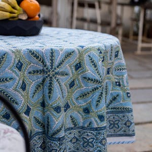 May include: A round tablecloth featuring a blue and green floral design. The tablecloth is draped over a round table. The pattern includes leaf and geometric shapes. The background includes a bowl of fruit.