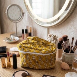 May include: A mustard yellow cosmetic bag with a white zipper and a floral pattern sits on a wooden vanity. Makeup brushes, lipsticks, and a compact are arranged around the bag, with a mirror in the background.