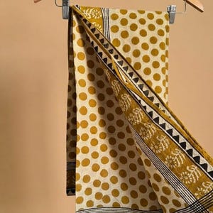 May include: A rectangular textile featuring a mustard yellow polka dot pattern on an off-white background. The fabric includes a decorative border with black triangular motifs, thin parallel lines, and a repeating floral print in mustard yellow.