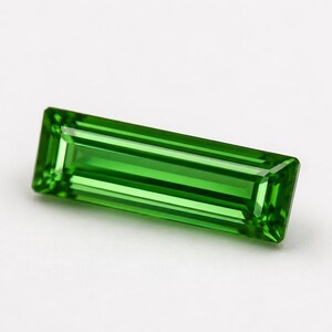 May include: A vibrant green gemstone cut into a long, rectangular baguette shape. The stone features precise, linear facets that create a clear, reflective surface with deep emerald tones and sharp, defined edges throughout the entire piece.