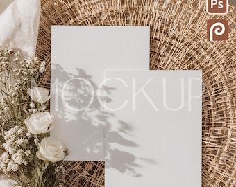 May include: A product mockup with two blank white cards, a bouquet of white flowers, and a woven straw placemat. The cards are arranged to display designs, with shadows adding depth. The scene is lit with soft, natural light.