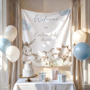 May include: A baby shower setup featuring a white cake, gifts, and balloons. A banner displays "Welcome to Emilias baby shower" with farm animal illustrations. The colour scheme is soft, with white, blue, and beige tones.