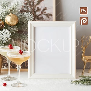 Puede incluir: Un marco de fotos blanco se encuentra sobre una superficie blanca, junto a dos copas de cóctel con cerezas. Una figura de ciervo dorado y ramas de árbol de Navidad están en el fondo. La escena tiene un tema festivo y navideño.