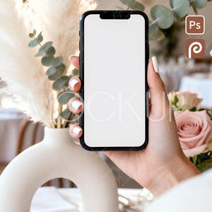 Può includere: Uno smartphone nero con schermo bianco vuoto è tenuto in una mano con unghie curate. Il telefono è posizionato davanti a uno sfondo sfocato con un vaso bianco, erba della pampa e fiori.