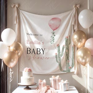 Pode incluir: Uma cena de ch&aacute; de beb&eacute; com uma faixa que diz "Welcome to Emilia's BABY shower". A faixa apresenta um cacto em aquarela e um bal&atilde;o rosa. Bal&otilde;es dourados, brancos e rosa decoram o quarto. Um bolo de dois andares e presentes embrulhados est&atilde;o em uma mesa.