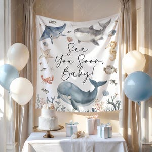 May include: A baby shower setup with a white cake, gifts, and balloons. A backdrop displays sea creatures and the text "Sea You Soon, Baby!". The colour scheme includes white, blue, and gold accents, creating a celebratory atmosphere.