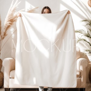 May include: A large, off-white blanket is held up in front of a person, obscuring their face. The blanket is the focal point, with a soft texture. The scene is set in a well-lit room with a neutral color palette, featuring a beige sofa and decorative plants.