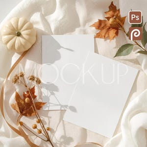 May include: A mockup scene featuring two blank white cards, a small white pumpkin, and autumn leaves. A gold ribbon and sprigs with berries add to the fall theme. The scene is set on a white, textured fabric.