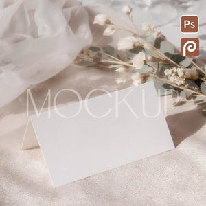 May include: A blank, white tent card stands on a textured, beige surface, with a soft, sheer fabric draped nearby. Delicate white flowers and greenery are arranged to the right, with a blurred background of glassware. The scene evokes a sense of elegance and is suitable for weddings or events.