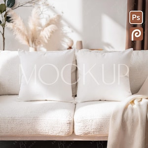 May include: Two white square pillows with the word "MOCKUP" printed on them, resting on a cream-colored textured sofa. A cream-colored blanket is draped over the sofa. The background includes decorative elements and a window with sunlight.