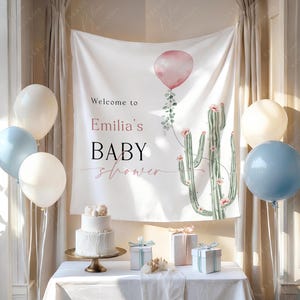 May include: A baby shower setup with a white banner displaying a pink balloon and cactus design, with the text "Welcome to Emilia's BABY shower." A two-tiered cake, gifts, and white and blue balloons are arranged on a table.