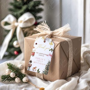 May include: A wrapped gift box in brown paper, tied with a natural twine ribbon and a decorative tag. The tag reads "Thank you for celebrating with us, Emilia's First Birthday." A small Christmas tree and decorative sprigs are in the background.