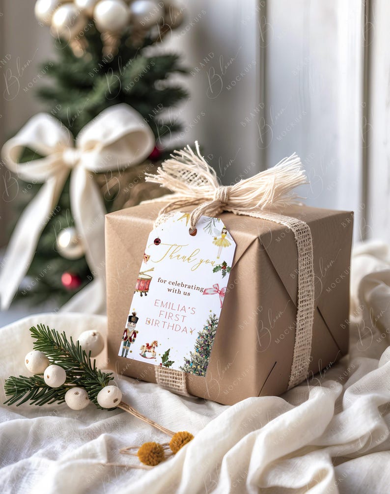 May include: A wrapped gift box in brown paper, tied with a natural twine ribbon and a decorative tag. The tag reads "Thank you for celebrating with us, Emilia's First Birthday." A small Christmas tree and decorative sprigs are in the background.