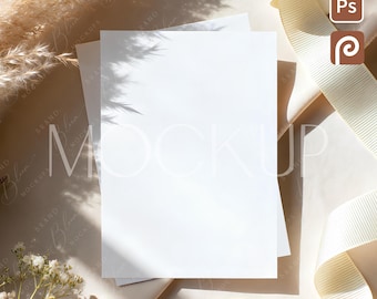 Puede incluir: Una maqueta con dos tarjetas blancas en blanco, una cinta color crema y flores secas. Las tarjetas están apiladas sobre una superficie beige, con sombras y luz solar. La imagen incluye el texto "MOCKUP" en una marca de agua.