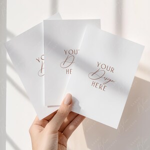 May include: A hand holding three white cards with the words "YOUR Design HERE" and "YOU' Design HERE" in a copper-colored script. The cards are blank, with a subtle texture, and are arranged in a fan shape against a soft, neutral background.