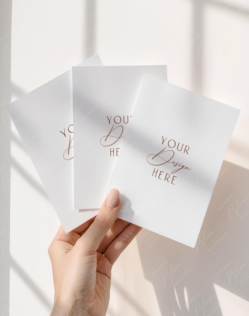 May include: A hand holding three white cards with the words "YOUR Design HERE" and "YOU' Design HERE" in a copper-colored script. The cards are blank, with a subtle texture, and are arranged in a fan shape against a soft, neutral background.