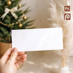 May include: A hand holding a blank white rectangular card, likely a mockup for a greeting card or invitation. A small Christmas tree with gold ornaments and a white bow is in the background, along with decorative pampas grass. The scene has a soft, neutral color palette.