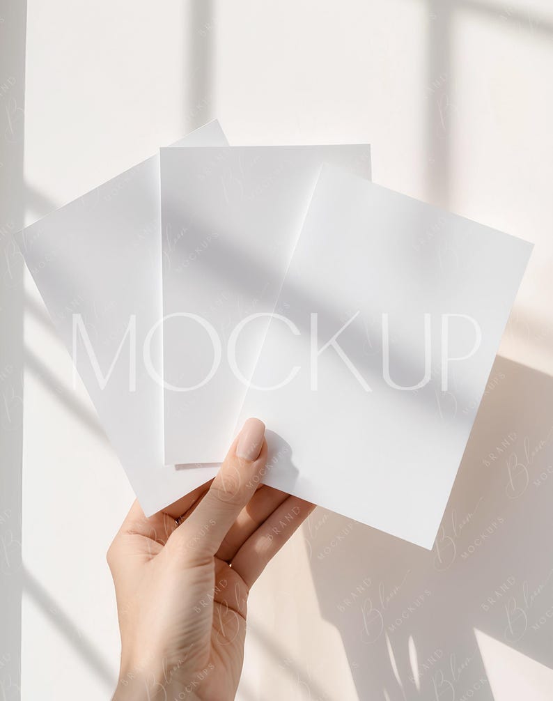 May include: A hand holding three blank white square cards, possibly invitations or stationery. The cards are positioned against a white background with soft shadows, creating a clean and minimalist aesthetic. The word "MOCKUP" is faintly visible.