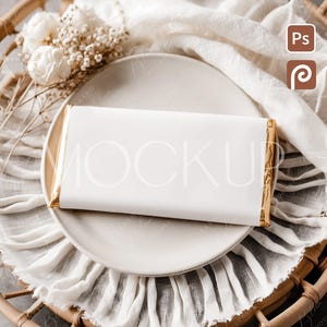 May include: A white chocolate bar with gold foil edges rests on a white plate, atop a textured white cloth. The plate sits on a woven wicker tray, with white flowers and dried stems. The image has a soft, neutral color palette.