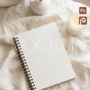 May include: A blank white spiral-bound notebook lies open on a cream-colored fabric surface. A small white vase with dried pampas grass and a lit candle in a glass jar are also present. The scene is a mockup, with the word "MOCKUP" faintly visible.
