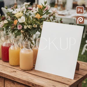 May include: A rustic wooden table displays a blank white sign, surrounded by glass bottles of juice in red, orange, and yellow hues. A vase of colorful flowers adds a decorative touch. The scene suggests a wedding or event setting.