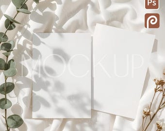 May include: Two white rectangular cards with the word "MOCKUP" printed on them, on a white, textured fabric. Green eucalyptus and small white flowers are arranged around the cards, casting shadows. The image has a soft, natural light.