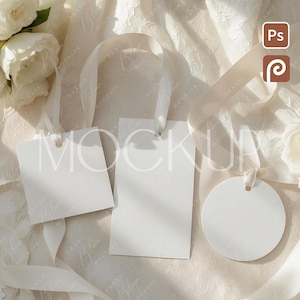 May include: Three blank white tags with attached ribbons, one square, one rectangular, and one circular, arranged on a textured, off-white surface. White roses and a flowing ribbon are also present, creating a soft, elegant aesthetic.