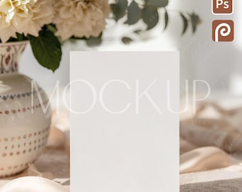 Puede incluir: Una tarjeta blanca en blanco se encuentra en posición vertical, centrada en una escena suave y soleada. Un jarrón de flores y vegetación color crema se encuentra a la izquierda, con una cinta y encaje que agregan textura. La palabra "MOCKUP" es visible.