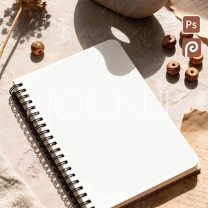 Puede incluir: Un cuaderno en espiral en blanco con una espiral negra, abierto en una página blanca. El cuaderno está sobre una superficie texturizada con cuentas de madera decorativas, flores secas y papel pergamino. La imagen tiene una estética neutra y terrosa.