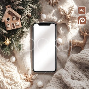 Może przedstawiać: Smartfon z pustym białym ekranem otoczony jest świątecznymi dekoracjami. Piernikowy domek, ozdoba w kształcie gwiazdy i złoty renifer ułożone są z białymi bombkami i przytulnym, dzianinowym kocem. Scena przywołuje świąteczny, zimowy motyw.