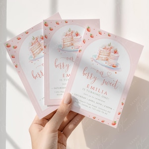 May include: Pink birthday invitations with a strawberry and cake design. The text reads "our berry sweet" and "Emilia is turning one!" with event details. The invitations are held in a hand.