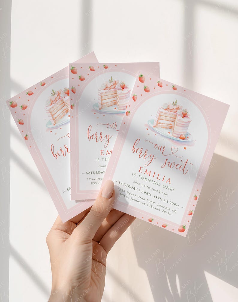 May include: Pink birthday invitations with a strawberry and cake design. The text reads "our berry sweet" and "Emilia is turning one!" with event details. The invitations are held in a hand.